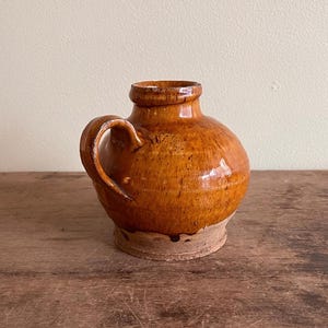 Può includere: Una piccola brocca rotonda in ceramica marrone con manico. La brocca ha una finitura lucida e una superficie strutturata. La base è non smaltata. La brocca è probabilmente vintage o antica.