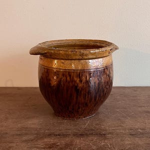 May include: A brown and tan ceramic pot with a wide rim and two small handles. The pot has a rounded body and a textured surface, with a darker brown base and lighter brown top.