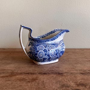 May include: A vintage blue and white ceramic creamer with a floral pattern. The creamer has a curved handle and a spout for easy pouring. The design features detailed floral motifs and a scalloped rim. The creamer is a classic piece of tableware.
