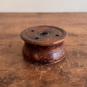 May include: A dark brown wooden spool with a small hole in the center and four smaller holes around the edge. The spool is sitting on a wooden surface.