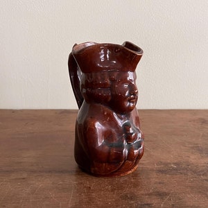 Pode incluir: Um jarro de cerâmica marrom em forma de pessoa usando um chapéu. O jarro tem um rosto detalhado e está segurando um pequeno objeto em sua mão.