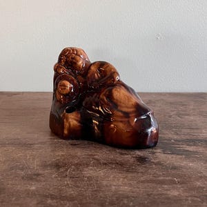 May include: A small, brown ceramic figurine with a glossy finish. The sculpture appears to depict a stylized, abstract form, possibly of a person or animal. The piece has a dark, wood-like appearance and sits on a wooden surface.