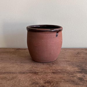 May include: A small, round, terracotta-colored ceramic pot with a dark brown rim. The pot has a slightly textured surface and two horizontal lines near the top. The interior is dark brown. The pot is likely used for planting or storage.