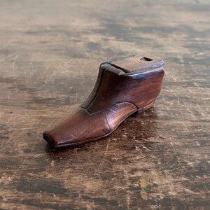 May include: A small, antique wooden shoe form in a rich, dark brown hue. The miniature shoe has a classic design with a defined toe, heel, and a small opening at the top. The wooden shoe form is displayed on a wooden surface.