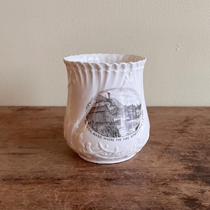 May include: White ceramic cup with a detailed relief pattern and a black and white image of a building and text that reads "Natick, Mass. Where the fire started 1651-1777". The cup is on a wooden surface.