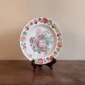 May include: A white ceramic plate with a floral design, featuring pink and red roses, green leaves, and red fruit. The plate has a scalloped edge and is displayed on a dark brown wooden stand.