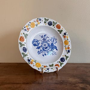 May include: A white ceramic plate with a blue floral design in the center and a decorative border of blue, orange, and yellow flowers. The plate is displayed on a white metal stand, showcasing its intricate details and vintage charm.