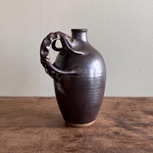 Puede incluir: Una jarra de cerámica marrón oscuro con un asa distintiva. La jarra tiene un cuerpo redondeado y un cuello estrecho. El asa está intrincadamente diseñada, agregando un toque único. La base de la jarra es de color marrón claro.