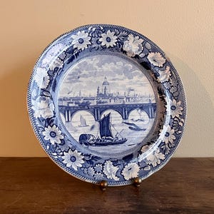 May include: A blue and white decorative plate with a detailed illustration of a bridge, boats, and a cityscape. The plate has a floral border and is displayed on a gold-coloured stand. The plate is a decorative item.