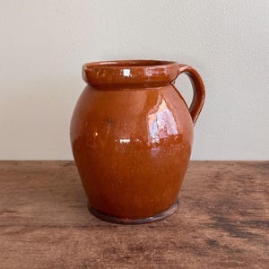 May include: A glazed, brown ceramic jug with a handle. The jug has a rounded body and a slightly flared rim. The surface reflects light, indicating a glossy finish. The jug is likely vintage or antique.