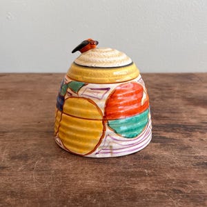 May include: A colorful ceramic jar with a lid, featuring a bird-shaped handle. The jar's body is adorned with yellow, orange, green, and blue geometric patterns. The lid is cream-colored with a yellow band. The jar sits on a wooden surface.