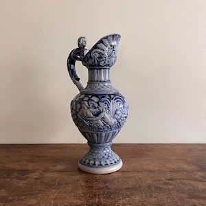 May include: A blue and white ceramic pitcher with a detailed relief design. The pitcher features a handle with a small figure, a decorative spout, and a bulbous body with a classical scene. The base is a pedestal design. The pitcher is on a wooden surface.