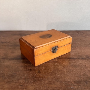 May include: A small wooden box with a hinged lid and a metal latch. The box is a light brown color and has a smooth finish.