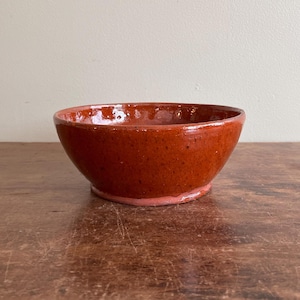 May include: A round, deep bowl with a glossy, dark reddish-brown glaze and a smooth, curved rim. The exterior surface features subtle speckling throughout the finish, and the base is slightly raised with a lighter, unglazed terracotta-colored edge.