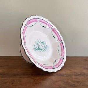 May include: A white ceramic bowl with a pink and green floral design. The bowl has a scalloped edge and is on a pedestal.