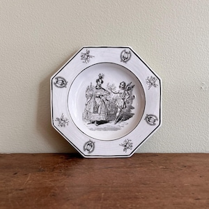 May include: White octagonal plate featuring a black and white illustration of two figures. The plate has a black border and decorative elements. It sits on a wooden surface.