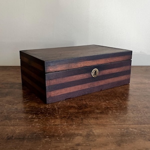 May include: A rectangular wooden box featuring alternating horizontal stripes of dark brown and reddish-brown wood. The box has a flat lid and a circular brass keyhole escutcheon centered on the front panel, showcasing a smooth, polished finish.