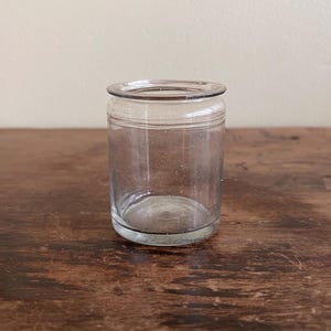 May include: A small, clear glass jar with a cylindrical shape. The jar has a slightly flared rim and a horizontal line etched around the middle. The glass is transparent, allowing a view of the interior. The jar sits on a dark wooden surface.