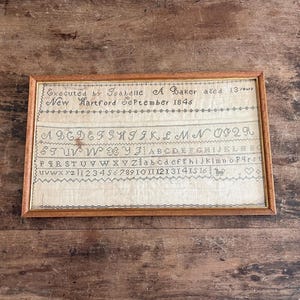 May include: Framed antique sampler with a wooden frame. The sampler, stitched on a light-colored fabric, features an alphabet, numbers, and the text "Executed by Isabelle A Baker aged 13 years New Hartford September 1846."
