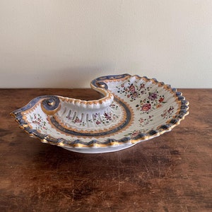 May include: A white porcelain serving dish with a scalloped edge and a floral design. The dish is shaped like a seashell and has a blue and gold trim.