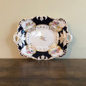 May include: An antique porcelain serving tray with a navy blue border and ornate gold trim. The tray features floral designs in pink, yellow, and blue, and has integrated handles. A decorative piece for serving or display.