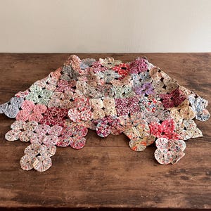 May include: A vintage, handmade textile piece composed of numerous fabric circles in various colors, including pink, red, and blue. The piece is laid on a dark wooden surface, showcasing its intricate design and craftsmanship. The textile has a scalloped edge.