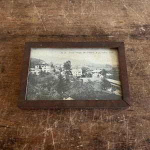 May include: A framed black and white photograph of a village scene, likely from the early 20th century. The frame is a dark brown wood. The photograph is of Shaker Village, Mt. Lebanon, N.Y., looking South.