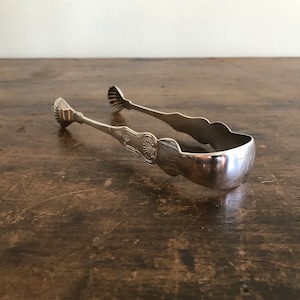 May include: Silver sugar tongs with an ornate design. The tongs are closed and lying on a wooden surface.