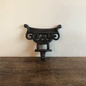 May include: A black cast iron decorative bracket with a scroll design. The bracket has a flat top and a pointed bottom.