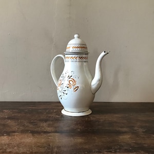 Puede incluir: Cafetera de cerámica blanca con tapa y diseño floral en naranja y negro. La cafetera tiene un pico y una manija.