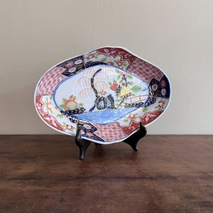May include: An oval-shaped ceramic serving dish with a white background and a colorful Japanese-inspired design. The design features a blue and white landscape with a willow tree, flowers, and a bridge. The dish is decorated with red, blue, gold, and black accents.