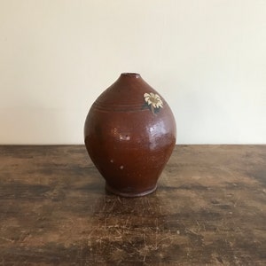 May include: A brown ceramic vase with a white flower painted on the side. The vase has a rounded body and a narrow neck.