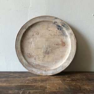 May include: A round wooden serving tray with a raised rim and a worn, whitewashed finish. The tray has a rustic, vintage look.