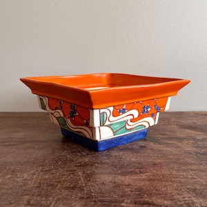 Puede incluir: Un cuenco cuadrado de cerámica con borde naranja y un diseño decorativo de diseños rojos, azules y verdes. El cuenco tiene una base blanca con fondo azul. El cuenco está sobre una superficie de madera.