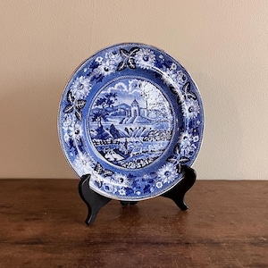 May include: A blue and white porcelain plate with a floral border and a central image of a landscape with a building. The plate is on a wooden stand.