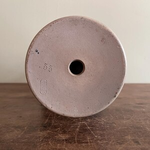 May include: A light brown ceramic object with a circular top and a central hole. The number ".55" is printed on the top, along with a rectangular symbol. The object is likely a vintage pottery piece.