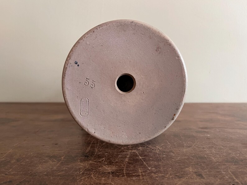 May include: A light brown ceramic object with a circular top and a central hole. The number ".55" is printed on the top, along with a rectangular symbol. The object is likely a vintage pottery piece.