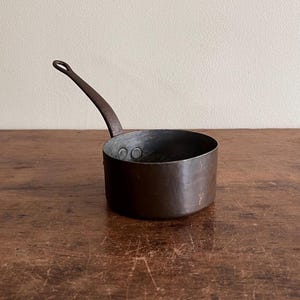 May include: A small copper saucepan with a long handle. The saucepan is on a wooden surface.