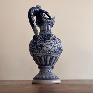 May include: A blue and white ceramic pitcher with a detailed relief design. The pitcher features a handle with a sculpted figure, and the body is adorned with a classical scene. The base is wide and the overall height is approximately 12 inches.