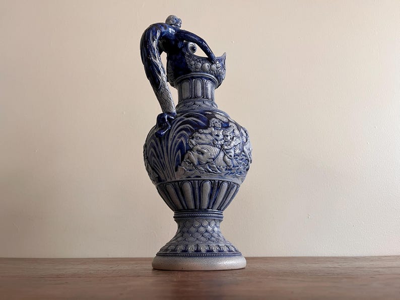 May include: A blue and white ceramic pitcher with a detailed relief design. The pitcher features a handle with a sculpted figure, and the body is adorned with a classical scene. The base is wide and the overall height is approximately 12 inches.