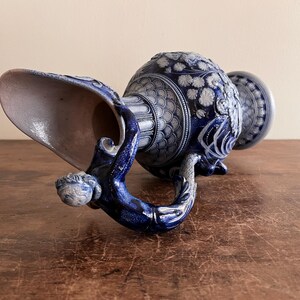 May include: A blue and white ceramic pitcher with intricate floral and geometric patterns. The handle is sculpted with a figure, and the spout is wide. The pitcher is resting on a wooden surface.