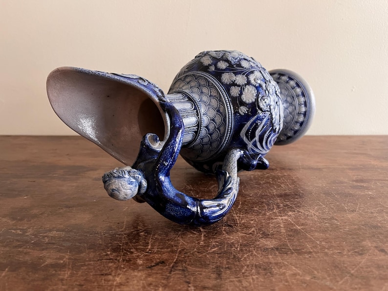 May include: A blue and white ceramic pitcher with intricate floral and geometric patterns. The handle is sculpted with a figure, and the spout is wide. The pitcher is resting on a wooden surface.