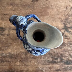May include: A blue and white ceramic pitcher with a detailed design. The pitcher has a handle and a spout, and the top is a light beige color. The pitcher is decorated with a floral pattern and a figure on the handle.