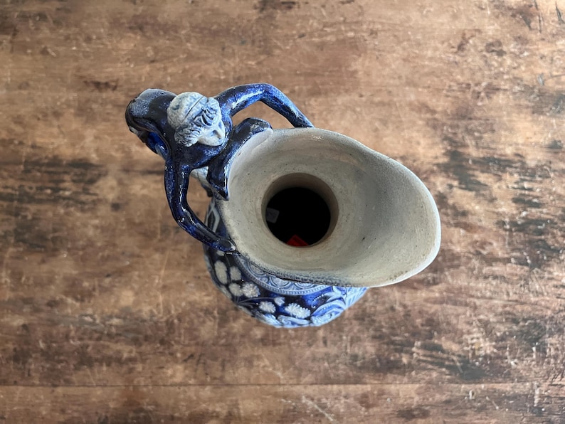 May include: A blue and white ceramic pitcher with a detailed design. The pitcher has a handle and a spout, and the top is a light beige color. The pitcher is decorated with a floral pattern and a figure on the handle.
