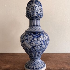May include: A blue and white ceramic vase with detailed relief carvings. The vase features a sculpted face at the top, a central section with figures, and a decorative base. The vase is displayed on a wooden surface.