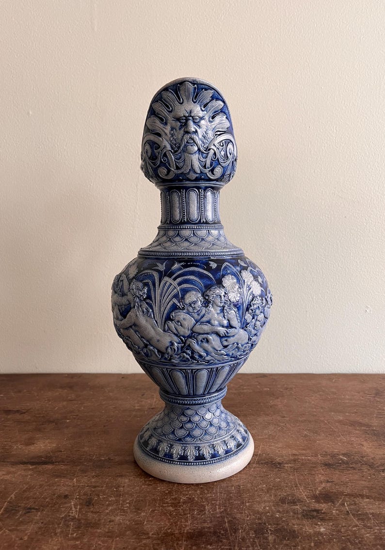 May include: A blue and white ceramic vase with detailed relief carvings. The vase features a sculpted face at the top, a central section with figures, and a decorative base. The vase is displayed on a wooden surface.