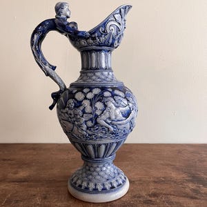 May include: A blue and white ceramic pitcher with a detailed relief design. The pitcher features a handle with a sculpted figure, and the body is adorned with classical figures and floral patterns. The pitcher stands on a pedestal base.