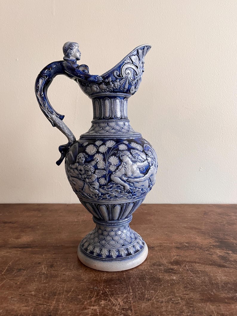 May include: A blue and white ceramic pitcher with a detailed relief design. The pitcher features a handle with a sculpted figure, and the body is adorned with classical figures and floral patterns. The pitcher stands on a pedestal base.