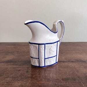 Puede incluir: Cremaera de cerámica blanca con ribetes azules y diseños en relieve. La cremaera tiene un cuerpo redondeado y un asa.