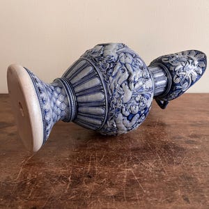 May include: A blue and white ceramic vase with intricate relief detailing. The vase features a wide base, a central body adorned with cherubic figures, and a spout with ornate patterns. The color scheme is a classic blue and white.